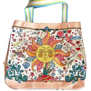 May include: A canvas tote bag with a whimsical design. The bag features a large sun illustration with a smiling face, surrounded by hearts, flowers, and nautical elements. The handles are striped with blue, yellow, and white. The bag has the text "Sing Your Own Special Song".