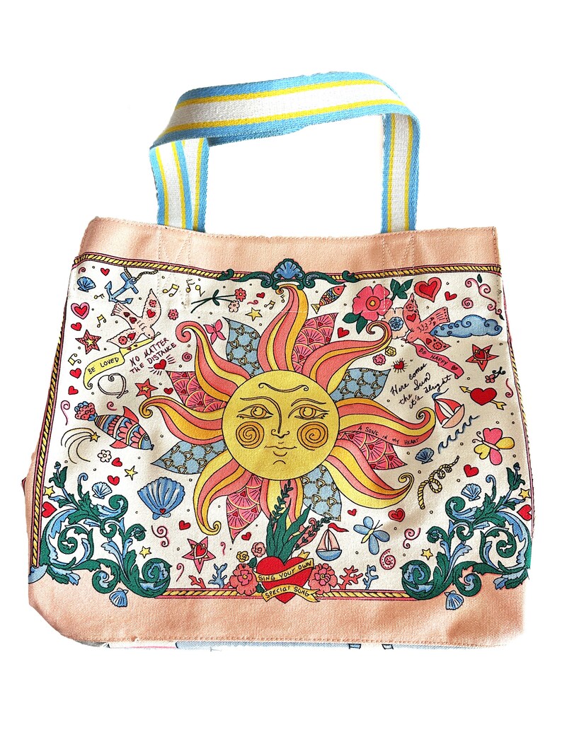 May include: A canvas tote bag with a whimsical design. The bag features a large sun illustration with a smiling face, surrounded by hearts, flowers, and nautical elements. The handles are striped with blue, yellow, and white. The bag has the text "Sing Your Own Special Song".