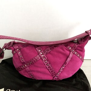 May include: A fuchsia-colored handbag with a curved shape. The bag features a suede body with crocodile-textured leather accents and silver-tone stud details. It has a matching strap and a zipper closure.