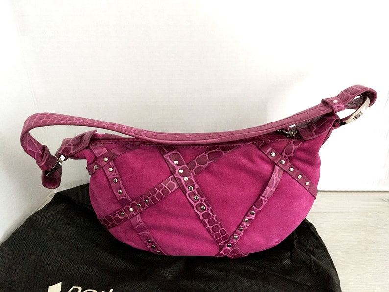 May include: A fuchsia-colored handbag with a curved shape. The bag features a suede body with crocodile-textured leather accents and silver-tone stud details. It has a matching strap and a zipper closure.