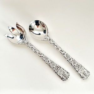 May include: A pair of ornate silver serving utensils, a spoon and a fork, with detailed floral and scrollwork patterns on the handles. The spoon has a rounded bowl, and the fork has three tines. The set is displayed on a white surface.
