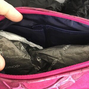 May include: A magenta-colored handbag with a textured exterior and a contrasting navy blue interior. The bag features a zippered closure and multiple interior pockets. The brand name "DONALD J PLINER" is visible on the packaging inside.