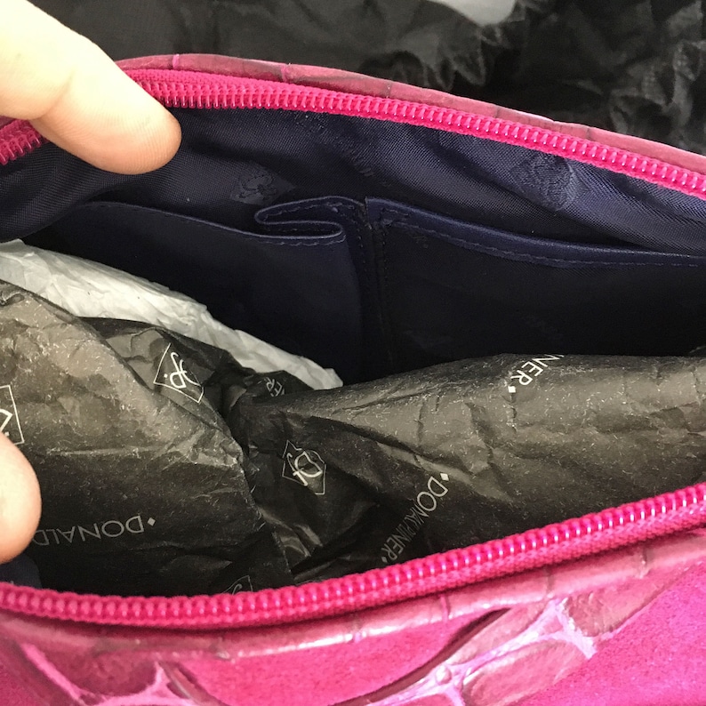 May include: A magenta-colored handbag with a textured exterior and a contrasting navy blue interior. The bag features a zippered closure and multiple interior pockets. The brand name "DONALD J PLINER" is visible on the packaging inside.