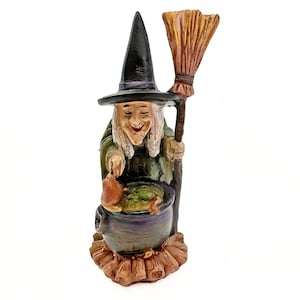 May include: A whimsical figurine of a smiling witch stirring a bubbling cauldron. She wears a black pointed hat, green robes, and holds a broom. The cauldron is purple, and the base is brown, creating a Halloween theme.