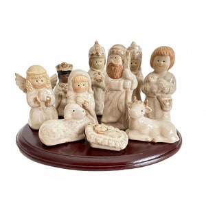 May include: A ceramic nativity set featuring ten glazed figurines in cream and brown tones, including an angel, a shepherd, a cow, a lamb, and a central manger scene, all arranged on a circular, dark brown wooden display base.