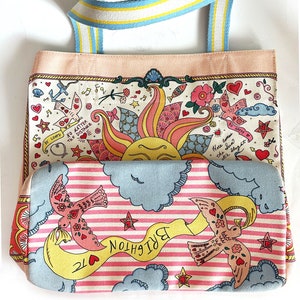 May include: A colorful tote bag with a whimsical design. The bag features a light pink base and a patterned fabric with illustrations of birds, hearts, and a sun. The handles are striped with blue, yellow, and white. Text includes "BE LOVED" and "BRIGHTON TC".
