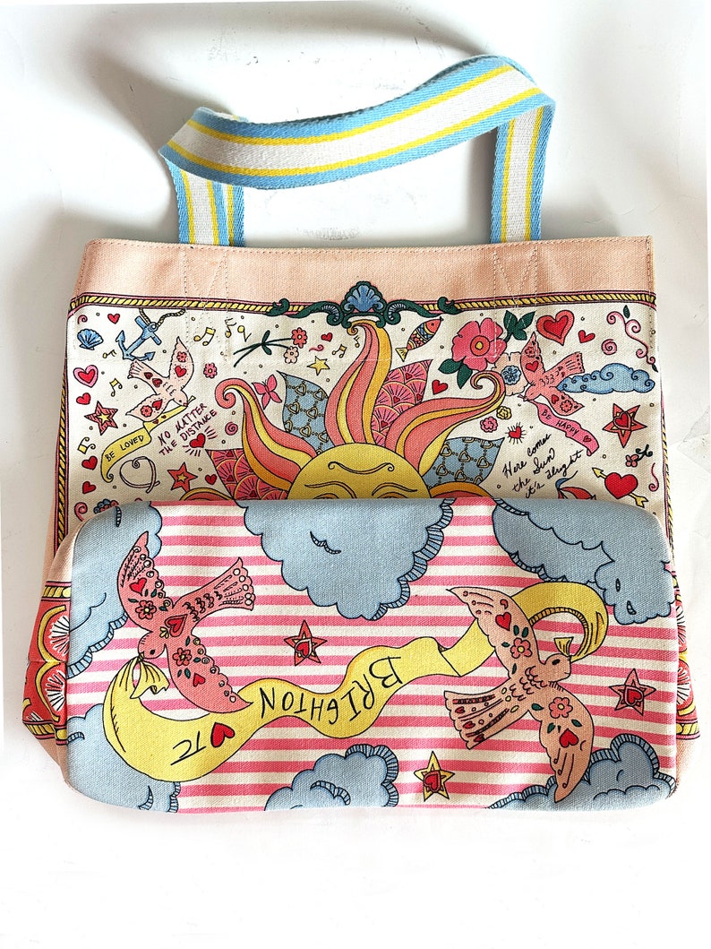 May include: A colorful tote bag with a whimsical design. The bag features a light pink base and a patterned fabric with illustrations of birds, hearts, and a sun. The handles are striped with blue, yellow, and white. Text includes "BE LOVED" and "BRIGHTON TC".
