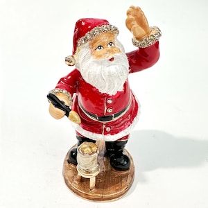 May include: A festive Santa Claus figurine in a red suit with a white beard and a red hat adorned with rhinestones. The figurine is holding a black object and standing on a wooden base, perfect for holiday decor.
