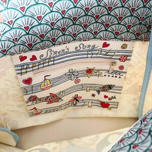 May include: A light blue and white tote bag with a seashell pattern and red heart accents. The interior features a zippered pocket with the text "Siren's Song" and nautical illustrations, including sailboats and musical notes.