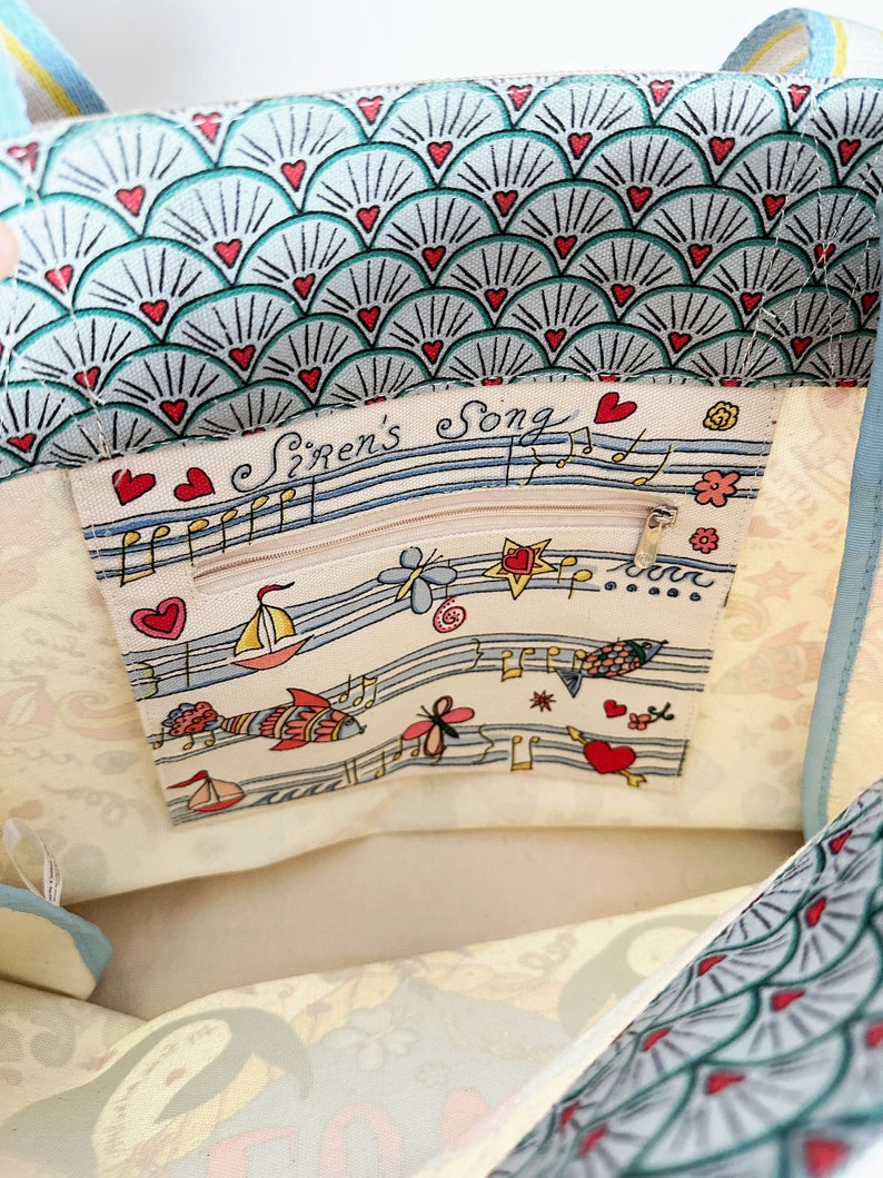 May include: A light blue and white tote bag with a seashell pattern and red heart accents. The interior features a zippered pocket with the text "Siren's Song" and nautical illustrations, including sailboats and musical notes.