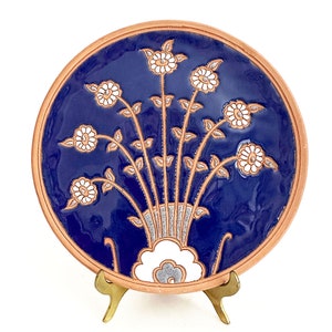 May include: A round decorative plate with a deep blue glazed surface featuring a hand-painted floral arrangement in white and light brown. The plate has a natural terracotta-colored rim and is supported by a small, gold-toned metal display stand.