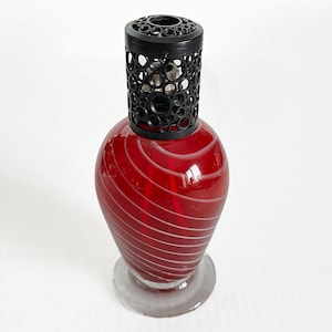 May include: A decorative red glass fragrance lamp with a black metal top. The lamp has a rounded body with white spiral stripes and a clear glass base. The black top has a circular pattern.