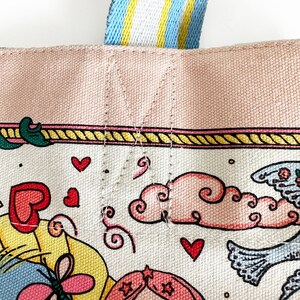 May include: Close-up of a canvas tote bag with a colorful illustration. The bag features a pink top border and a striped blue, yellow, and white handle. The illustration includes hearts, clouds, and a woman with pink hair.