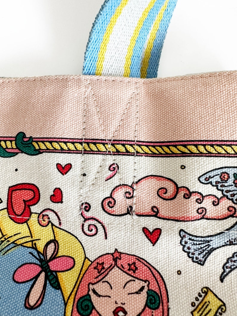 May include: Close-up of a canvas tote bag with a colorful illustration. The bag features a pink top border and a striped blue, yellow, and white handle. The illustration includes hearts, clouds, and a woman with pink hair.