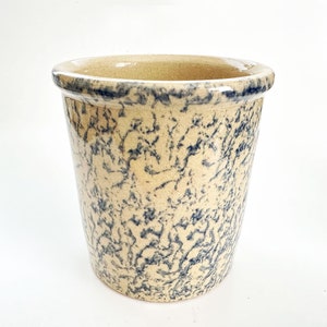 May include: A cylindrical stoneware crock with a wide opening. The exterior features a beige background with a dark blue, speckled pattern. The rim is dark blue. This vintage-style container is suitable for kitchen or home decor.