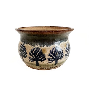 May include: A handcrafted ceramic bowl with a wide, rounded shape. The bowl features a dark brown rim and a design of stylized blue trees against a beige and green background. The bowl is suitable for decorative or functional use.