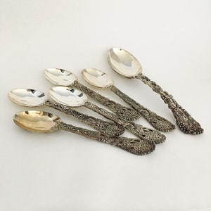 May include: Six silver spoons with ornate, floral-patterned handles. The spoons have a shiny, reflective surface and are arranged diagonally across a white background. The handles feature a detailed design of vines and leaves.