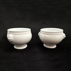 May include: Two white ceramic bowls with a lion's head design on the sides. The bowls have a rounded shape and sit on a small, circular base. The background is a solid black.