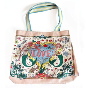 May include: A canvas tote bag with a whimsical mermaid design. The bag features a light pink border and a blue, yellow, and white striped handle. The main design includes mermaids, hearts, flowers, and the word "LOVE".