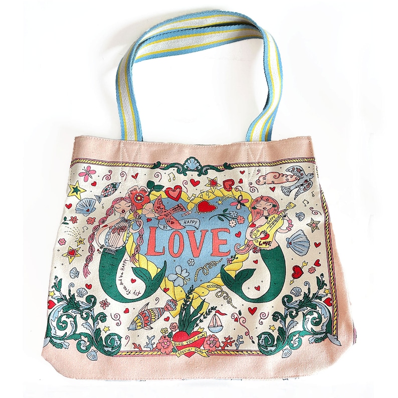 May include: A canvas tote bag with a whimsical mermaid design. The bag features a light pink border and a blue, yellow, and white striped handle. The main design includes mermaids, hearts, flowers, and the word "LOVE".