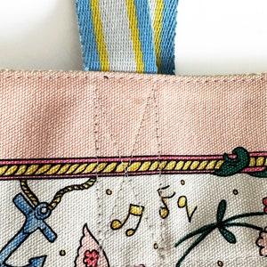 May include: Close-up of a canvas tote bag with a light pink top and a white base decorated with colorful illustrations. The bag has a blue, yellow, and white striped handle. The illustrations include an anchor, musical notes, and flowers.