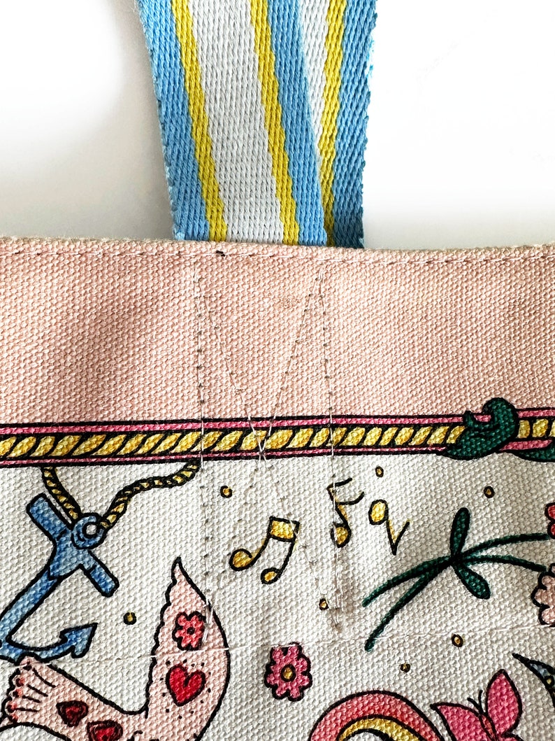 May include: Close-up of a canvas tote bag with a light pink top and a white base decorated with colorful illustrations. The bag has a blue, yellow, and white striped handle. The illustrations include an anchor, musical notes, and flowers.