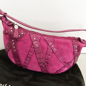 May include: A magenta suede shoulder bag with crocodile-textured leather accents and silver-tone hardware. The bag features a curved shape, a long strap, and decorative straps with silver studs. A black dust bag is visible below the purse.