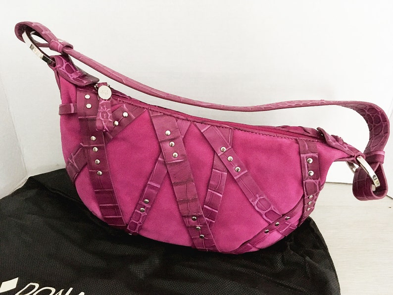 May include: A magenta suede shoulder bag with crocodile-textured leather accents and silver-tone hardware. The bag features a curved shape, a long strap, and decorative straps with silver studs. A black dust bag is visible below the purse.