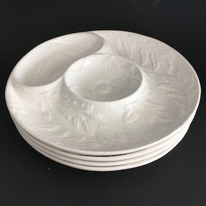 May include: A stack of four white ceramic serving dishes. Each dish has a central circular well and two side compartments. The dishes feature a raised floral and leaf pattern, and are stacked on a dark surface.