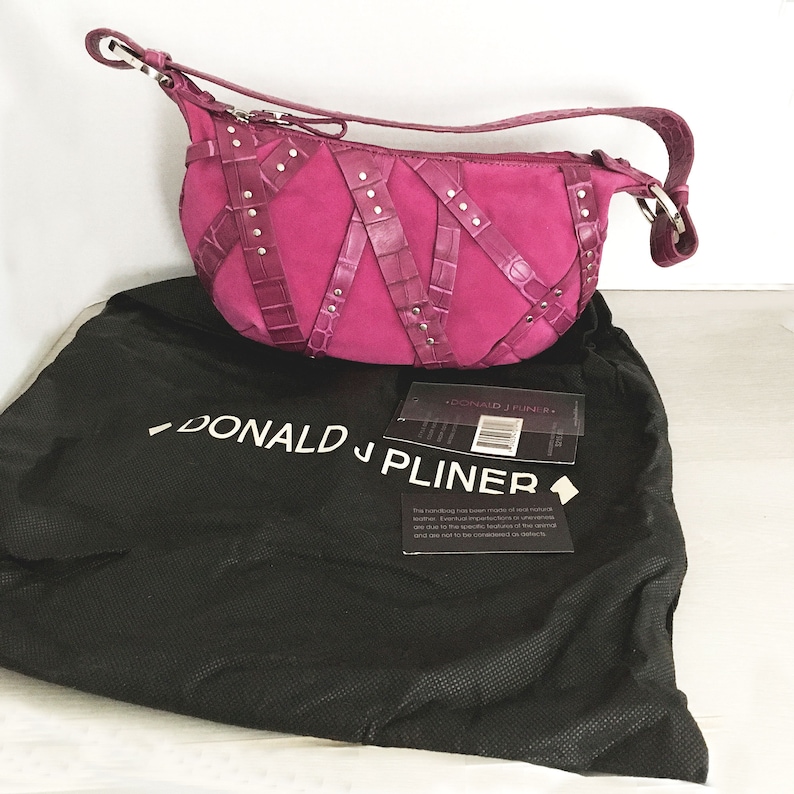 May include: A magenta suede handbag with crocodile-textured leather accents and silver-tone studs. The bag has a shoulder strap and a zipper closure. It rests on a black dust bag with the brand name "DONALD J PLINER" printed in white.
