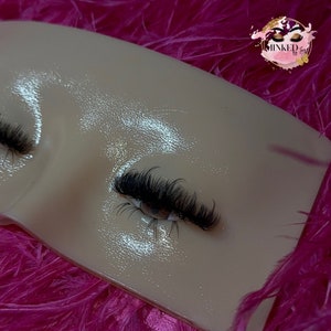 Könnte beinhalten: Nahaufnahme eines Wimpern-Übungsmodells mit langen, dunklen Wimpern, die auf das obere Augenlid aufgetragen wurden. Das Modell besteht aus einem hellfarbigen, strukturierten Material.