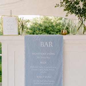 May include: A light blue bar menu with a frayed edge, listing drinks like The Daisy-Rita, beer, white and red wines. A gold framed sign says "Cheers" and a wedding date. The bar is decorated with flowers and glassware.