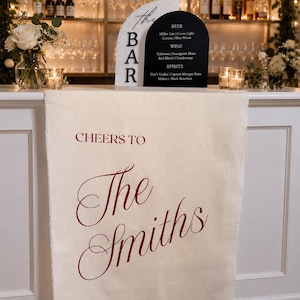 May include: A bar setup with a white bar, floral arrangements, and a sign listing beer, wine, and spirits. A cream-colored banner reads "Cheers to The Smiths" in burgundy script. The bar has a black and white sign that says "The BAR".