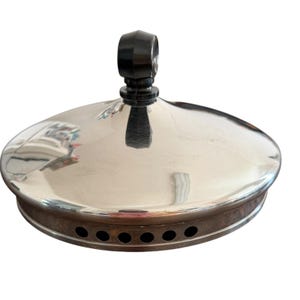 May include: A circular, polished silver-tone metal lid featuring a smooth, reflective domed surface and a central black knob handle. The lower edge of the lid is finished with a band containing a series of small, evenly spaced circular ventilation holes.