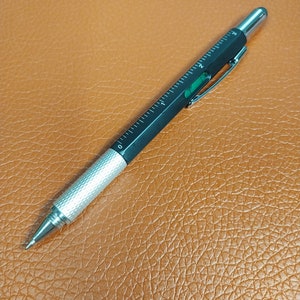 May include: A black and silver multi-tool pen with a ruler, level, and stylus. The pen is on a brown leather surface.
