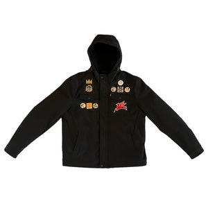 Puede incluir: Una chaqueta negra con capucha, cierre de cremallera y varios parches, entre ellos un caballo alado rojo, una corona y varios dise&ntilde;os m&aacute;s.