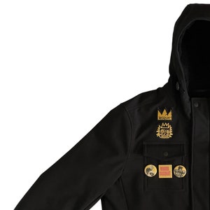 Puede incluir: Chaqueta negra con capucha con varios parches, incluyendo una corona con el texto "Basquiat" y un gr&aacute;fico amarillo y negro con el texto "Day", y dos parches redondos con el texto "Warhol Basquiat".