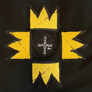 Puede incluir: Un parche de tela negro con un dise&ntilde;o bordado amarillo. El dise&ntilde;o presenta una gran estrella de cuatro puntas con un c&iacute;rculo m&aacute;s peque&ntilde;o en el centro. El c&iacute;rculo tiene el texto "SAMOWAS" escrito en blanco.