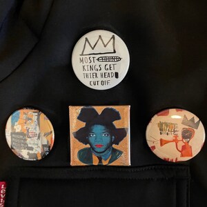 Puede incluir: Cuatro pines redondos con diferentes dise&ntilde;os. Un pin es blanco con texto negro que dice "Most Kings Get Their Head Cut Off." Otro pin presenta una ilustraci&oacute;n colorida de una persona con una corona. El tercer pin es una ilustraci&oacute;n colorida de una persona tocando la trompeta. El cuarto pin es una ilustraci&oacute;n colorida de una persona con una corona.