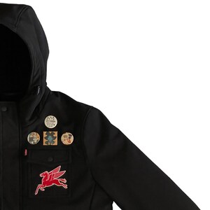 Puede incluir: Chaqueta softshell negra con cierre de cremallera y capucha. La chaqueta tiene varios parches y pines, incluido un logotipo de caballo rojo, un dise&ntilde;o floral y una corona.