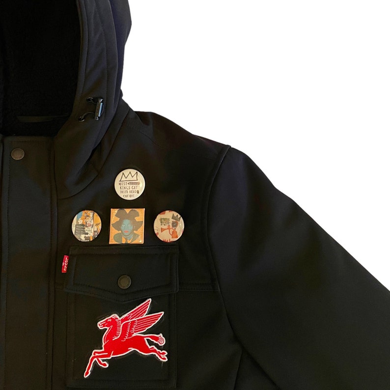 Puede incluir: Chaqueta negra con un parche de caballo alado rojo y varios pines redondos peque&ntilde;os. Los pines muestran una corona, una persona con cabello azul y una persona tocando la trompeta. El texto en uno de los pines dice "Wills Kingdom Kings Get Their Heads Cut Off".