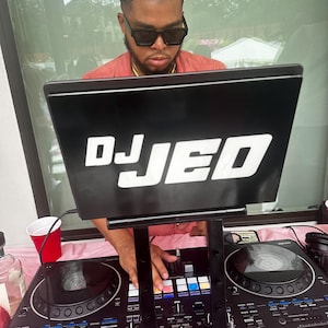 May include: A DJ with a black laptop that says "DJ JED" on the screen, is mixing music on a black DJ mixer with two turntables. The DJ is wearing a pink shirt and sunglasses.