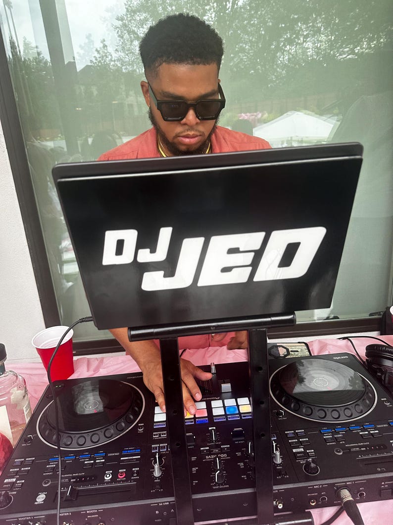May include: A DJ with a black laptop that says "DJ JED" on the screen, is mixing music on a black DJ mixer with two turntables. The DJ is wearing a pink shirt and sunglasses.