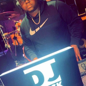 May include: A person wearing a black hoodie with a white Nike swoosh logo and a gold chain necklace is standing behind a DJ mixer. The mixer has a black surface with white text that reads "DJ BLACK" and "DJ EXPERIENCE".