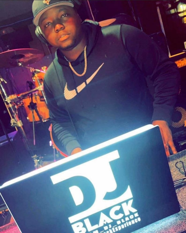 May include: A person wearing a black hoodie with a white Nike swoosh logo and a gold chain necklace is standing behind a DJ mixer. The mixer has a black surface with white text that reads "DJ BLACK" and "DJ EXPERIENCE".