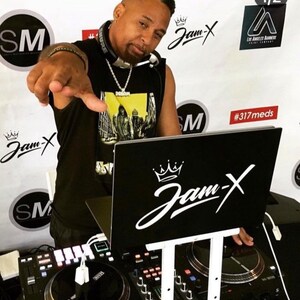 May include: A man wearing a black tank top with a yellow graphic print and a silver chain necklace is standing behind a DJ mixer. The mixer has two turntables and a control panel. The man is pointing at the camera with his right hand. The background is white with black and white text that says "Jam-X" and "#317meds".