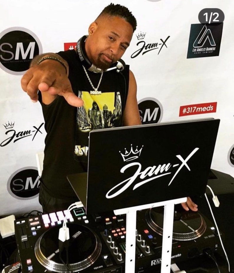 May include: A man wearing a black tank top with a yellow graphic print and a silver chain necklace is standing behind a DJ mixer. The mixer has two turntables and a control panel. The man is pointing at the camera with his right hand. The background is white with black and white text that says "Jam-X" and "#317meds".
