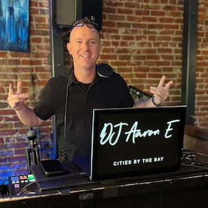 May include: A DJ with a black shirt and glasses is standing behind a DJ booth with a black and white sign that says "DJ Aaron E Cities by the Bay".