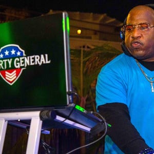 May include: A DJ wearing a blue shirt and headphones, standing behind a laptop with a green screen that says "PARTY GENERAL" in white text with a red, white, and blue star design.