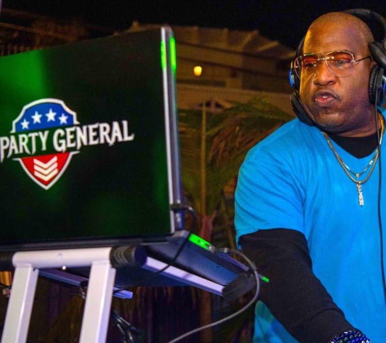 May include: A DJ wearing a blue shirt and headphones, standing behind a laptop with a green screen that says "PARTY GENERAL" in white text with a red, white, and blue star design.